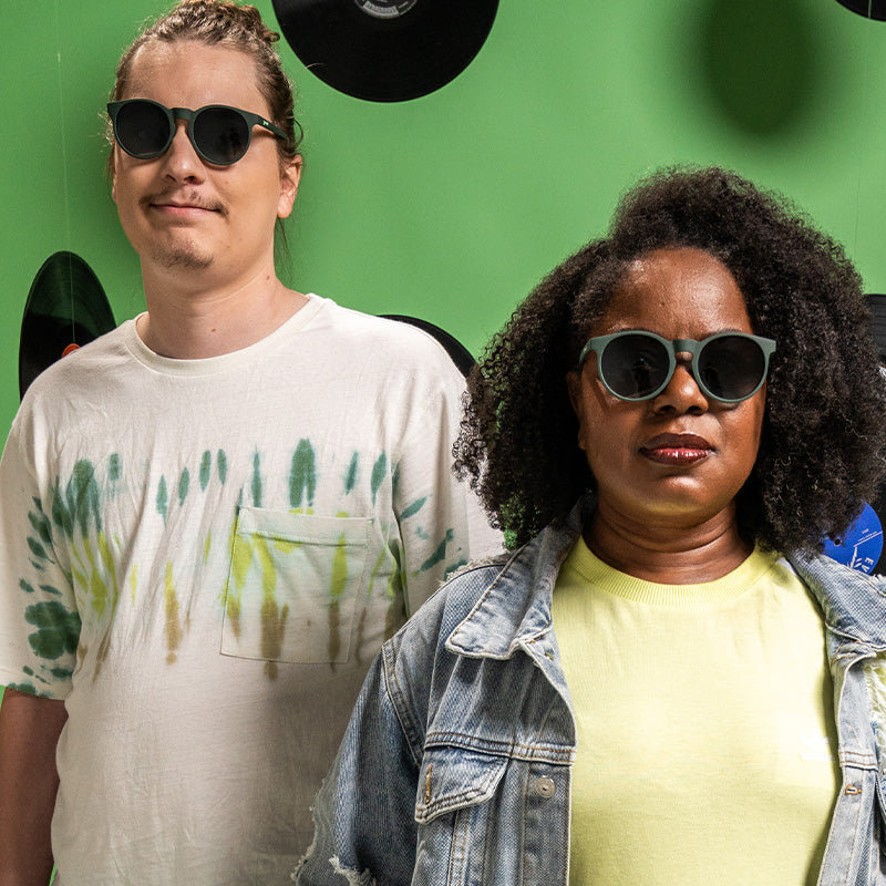 A man and woman wearing round dark green sunglasses with round green lenses look ahead, vinyl records behind them.